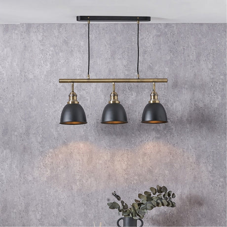Augusto Black & Gold Task Bar Pendant Light with three matt black domed shades on a brass bar and ceiling plate, shown against a grey textured wall.