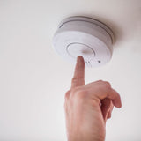 Finger pressing the hush button on the AVSL Mercury white photoelectric smoke detector mounted to a ceiling, showing the moulded test and warning labels.