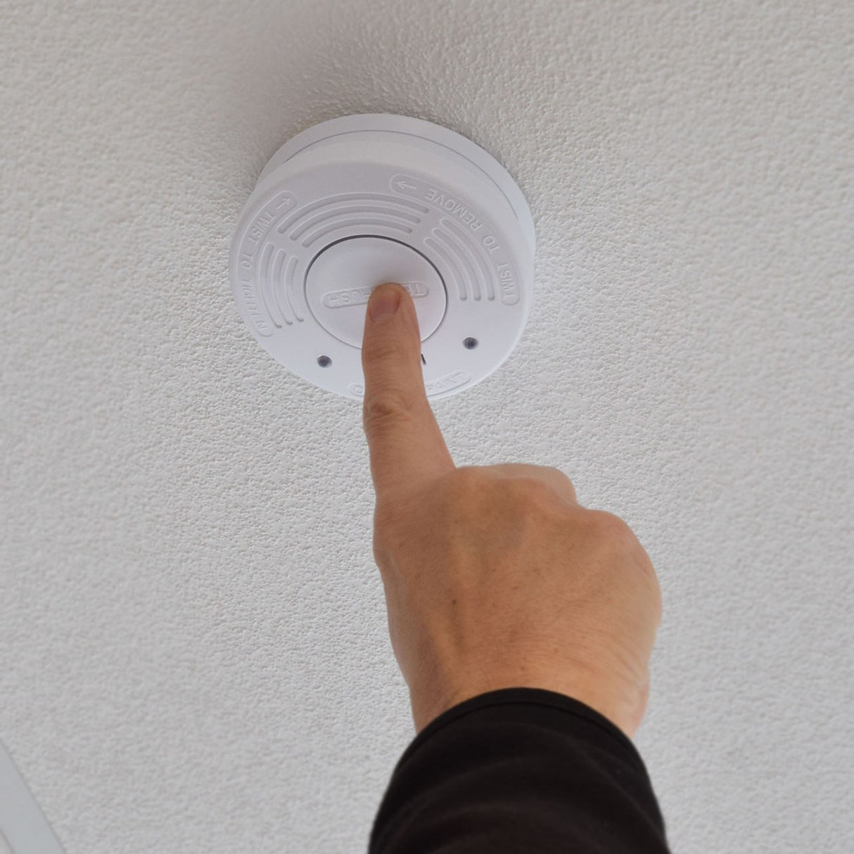 Finger pressing the central test/hush button on a ceiling-mounted Mercury photoelectric smoke detector with white ABS housing and twist-to-remove markings.