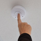 Finger pressing the central test/hush button on a ceiling-mounted Mercury photoelectric smoke detector with white ABS housing and twist-to-remove markings.