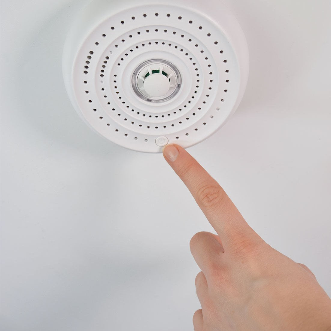 Finger pressing the test button on AVSL Mercury Heat Alarm, a round white ceiling-mounted unit with perforated cover and central thermistor sensor.