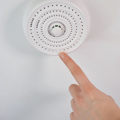 Finger pressing the test button on AVSL Mercury Heat Alarm, a round white ceiling-mounted unit with perforated cover and central thermistor sensor.