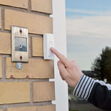 Finger pressing the white Mercury wireless doorbell transmitter mounted on a brick wall beside an older intercom unit.