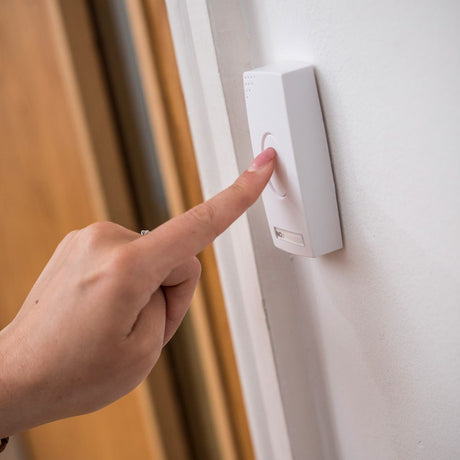 Finger pressing the white waterproof push button transmitter of the AVSL Mercury DB300 Wireless Door Chime, mounted on a door frame by a white wall.