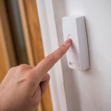 Finger pressing the white waterproof push button transmitter of the AVSL Mercury DB300 Wireless Door Chime, mounted on a white door frame.