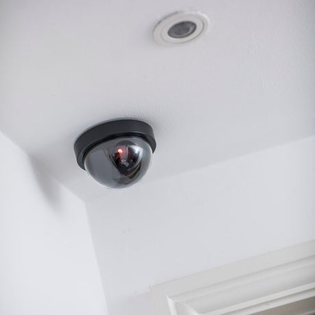 AVSL Mercury dummy dome CCTV camera in black with translucent dome, flashing red LED and dark grey base, ceiling-mounted in the corner of a white room.