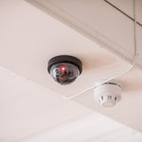 AVSL Mercury dummy dome CCTV camera in black with translucent dome and flashing red LED, ceiling-mounted beside a white smoke detector.