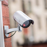 AVSL Mercury dummy IR bullet CCTV camera in white moulded plastic with flashing red LED and adjustable wall bracket, mounted on a red brick wall.