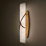 Valora LED wall light with a vertical alabaster column wrapped by curved brushed brass bands, illuminated warm white, mounted on a dark wall.