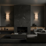 A modern living room with a dark marble fireplace, Glorina LED Small Stacked Alabaster Wall Lights – Soft White on either side, minimalist shelves with vases, a low coffee table, and a gray sectional sofa in moody lighting.