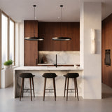 Modern kitchen with dark wood cabinets, a marble island with three black barstools, pendant lights, built-in oven, and large windows. The Vera LED Small Silhouette Alabaster Wall Light – Soft White adds subtle elegance above the potted plant.