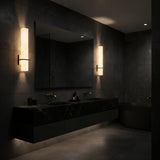 A modern, dark-toned bathroom with black marble sinks, a large mirror, a black bathtub, and Aurevon LED Vertical Panel Alabaster Wall Lights in soft white with brushed brass, all bathed in gentle ambient lighting.