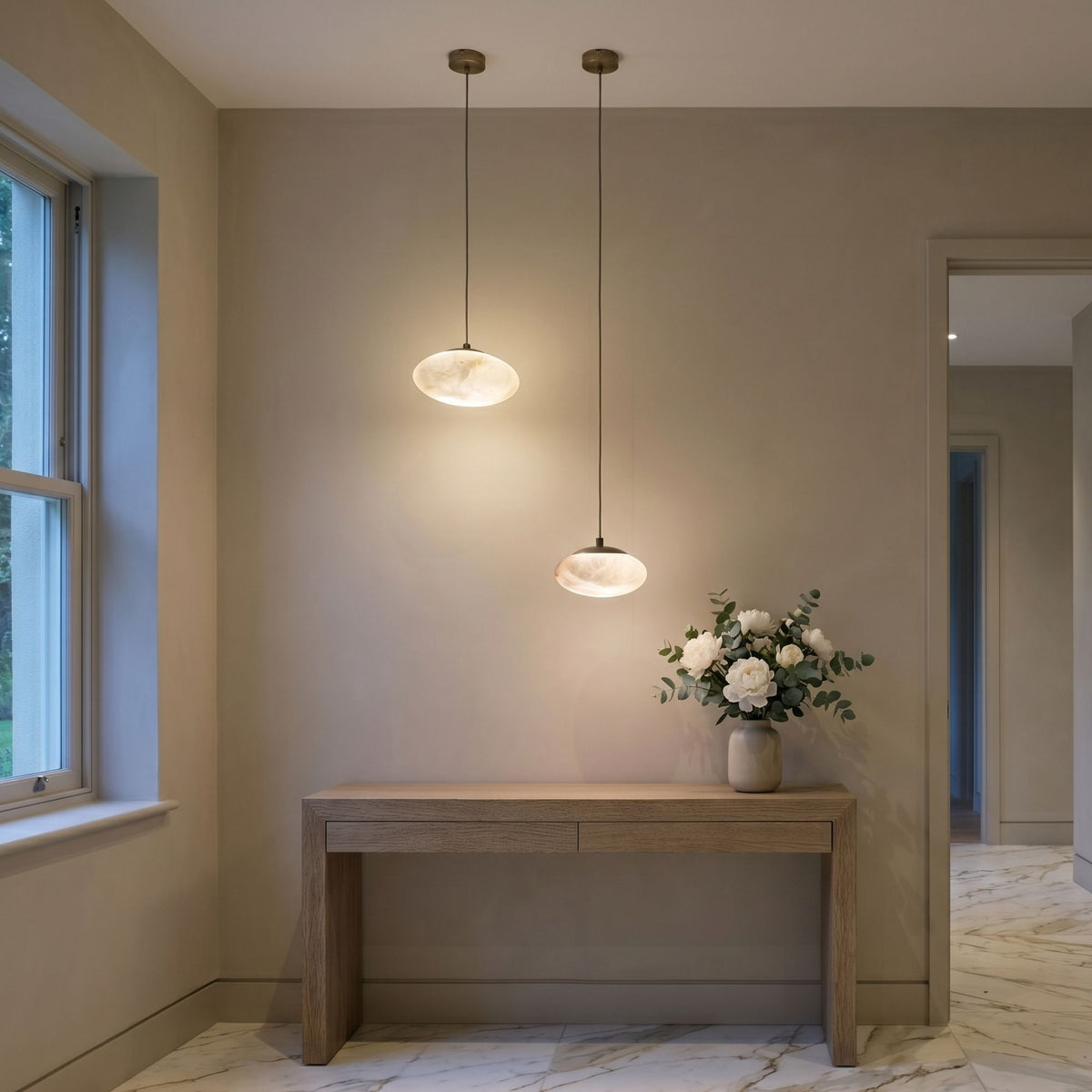 Two Solenne alabaster orb pendant lights with matt black caps hang above a wooden console table in a hallway with marble flooring.