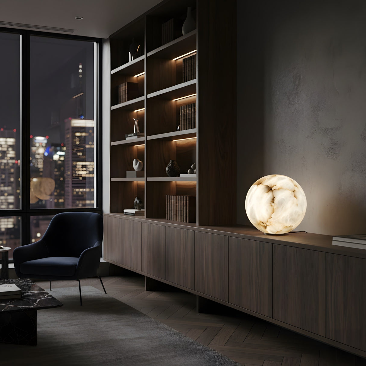 A modern living room with a built-in wooden bookshelf, a dark armchair, large windows revealing the city skyline at night, and a Zavero 1 Light Orb Alabaster Table Lamp - Soft White glowing on a cabinet.
