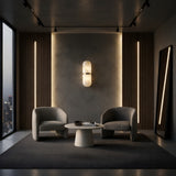 A modern lounge with two armchairs, a round coffee table, and a wall adorned with Ciel LED Cylinder Alabaster Wall Lights in Soft White & Brass. Large windows reveal a city view, and a tall mirror leans stylishly against the wall.