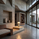 Modern living room with floor-to-ceiling windows, concrete walls and floor, built-in wooden shelves displaying ceramics, a minimalist wooden sofa, and a table with the Ascot 4 Light Small Table Lamp in White & Gold. Natural light fills the space.