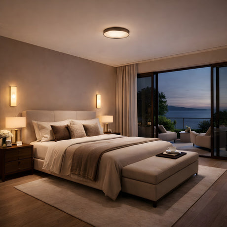 A modern bedroom featuring a large bed with neutral bedding, matte black nightstands, soft lighting from the Marea Round LED Alabaster Semi-Flush Ceiling Light in Matte Black, and a sliding glass door to a balcony overlooking a sunset on the water.