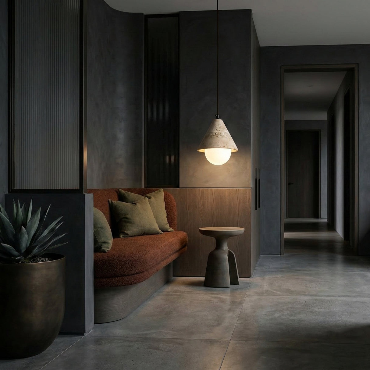 A modern, minimalist interior with a dark palette, featuring a brown cushioned bench with green pillows, a small round side table, the Alvero 1 Light Cone Travertine Single Pendant Light in light beige, and a potted plant on polished concrete.