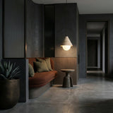 A modern, minimalist interior with a dark palette, featuring a brown cushioned bench with green pillows, a small round side table, the Alvero 1 Light Cone Travertine Single Pendant Light in light beige, and a potted plant on polished concrete.