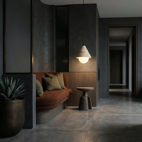 A modern, minimalist interior with a dark palette, featuring a brown cushioned bench with green pillows, a small round side table, the Alvero 1 Light Cone Travertine Single Pendant Light in light beige, and a potted plant on polished concrete.