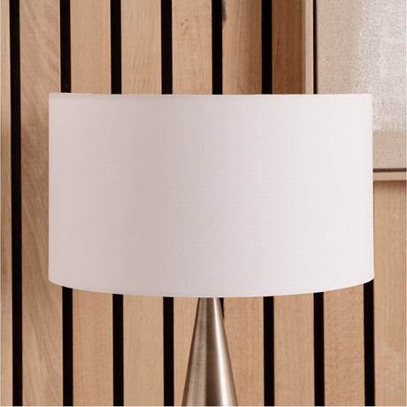Harry Ivory Cotton Mix Cylinder Lampshade, 25cm, shown fitted on a table lamp with brushed metal base against a light wood slatted wall.