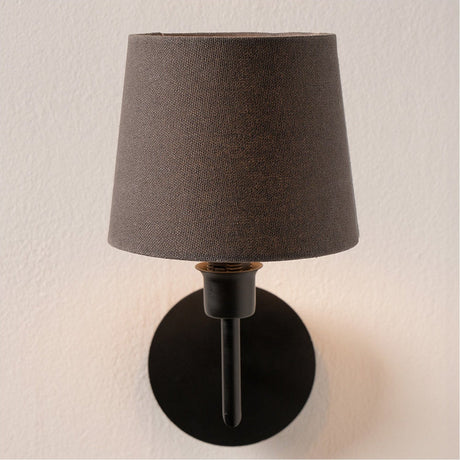 Pelham Grey Handloom Tapered Candle Clip Lampshade in grey fabric, shown fitted on a matte black wall light with a candle-style fitting against a plain wall.