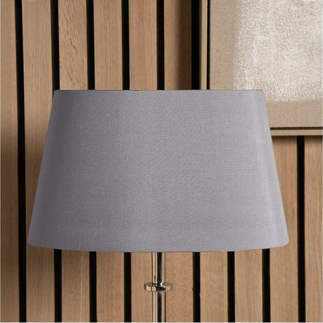 Martha Steel Grey Oval Silk Mix Tapered Lampshade, 25cm, shown fitted on a lamp base with a slatted wood panel background.