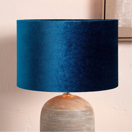 Bow Sapphire Velvet Cylinder Lampshade, 25cm, in deep blue velvet on a table lamp with a rounded ceramic base, set against a soft pink wall.