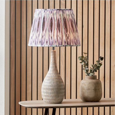 Izara Lilac Ikat Pleated Tapered Lampshade, 30cm, in fabric with a purple patterned print, shown fitted on a textured ceramic table lamp base.