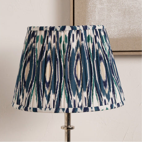 Izara Ocean Blue Ikat pleated tapered lampshade, 30cm, in a fabric print of navy, teal and cream, shown fitted to a chrome lamp base against a neutral wall.