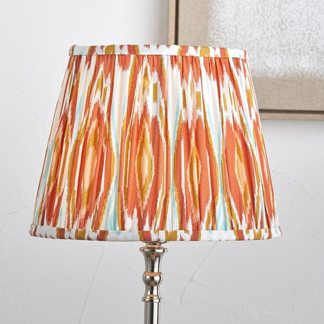 Izara Cinnamon Ikat Pleated Tapered Lampshade, 35cm, in fabric with orange, gold and white patterned design, shown on a chrome table lamp base.