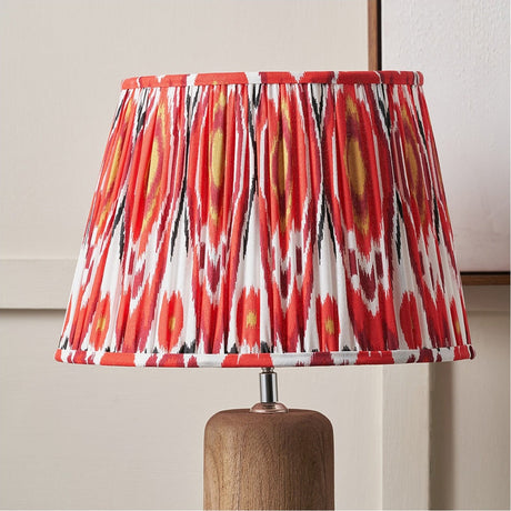 Izara Red Ikat Pleated Tapered Lampshade, 35cm, in red, black and yellow ikat pattern fabric, shown on a wooden lamp base against a neutral wall.