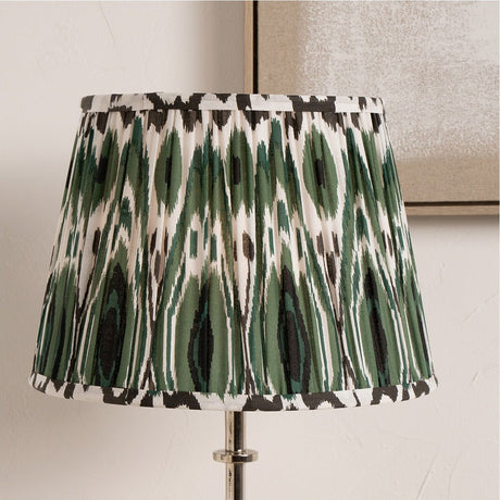 Izara Racing Green Ikat Pleated Tapered Lampshade, 35cm, on a chrome table lamp base, pleated fabric in green, black and cream ikat pattern, set against a light neutral wall.