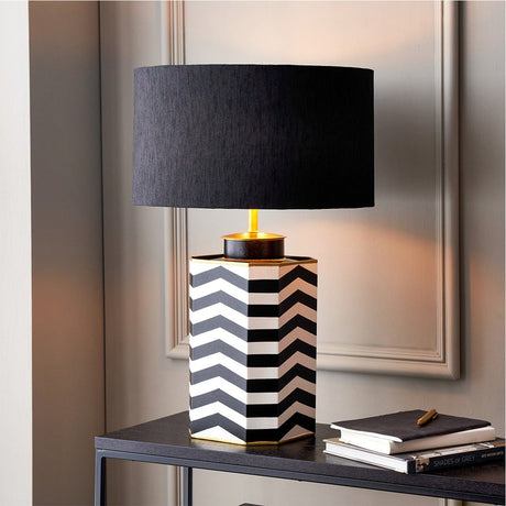 Stellan black slubbed silk cylinder lampshade, 45cm, with gold-lined interior, shown on a chevron-patterned lamp base with gold fitting in a living room setting.