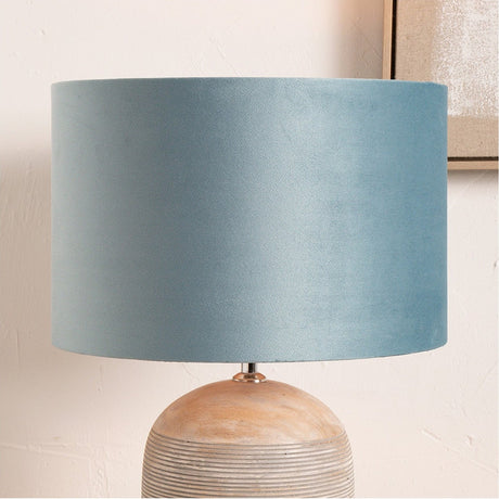 Rene Duck Egg Velvet Cylinder Lampshade, 45cm wide, in soft blue fabric on a lamp with a rounded wooden base, set against a light interior wall.