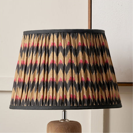 Athira Multi Chevron Design Pleated Tapered Lamp Shade, 30cm, in gold, pink and black zigzag fabric with dark trim, shown on a wooden table lamp base.