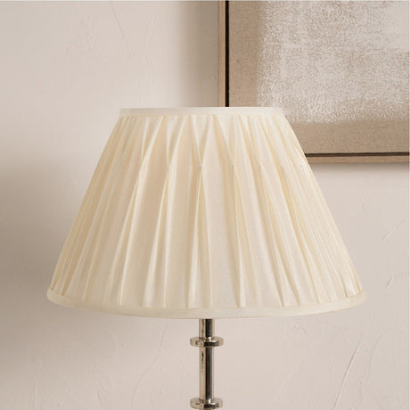 Garbo Cream Silk Mix Pinch Pleated Tapered Lampshade, 45cm, shown fitted on a chrome table lamp base against a plain wall with a framed artwork nearby.