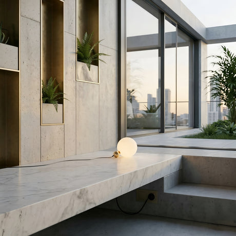 Modern interior with concrete walls, built-in plants, a glowing Soul Table Lamp - Gold on a marble ledge, large glass windows, and cityscape views at sunset.