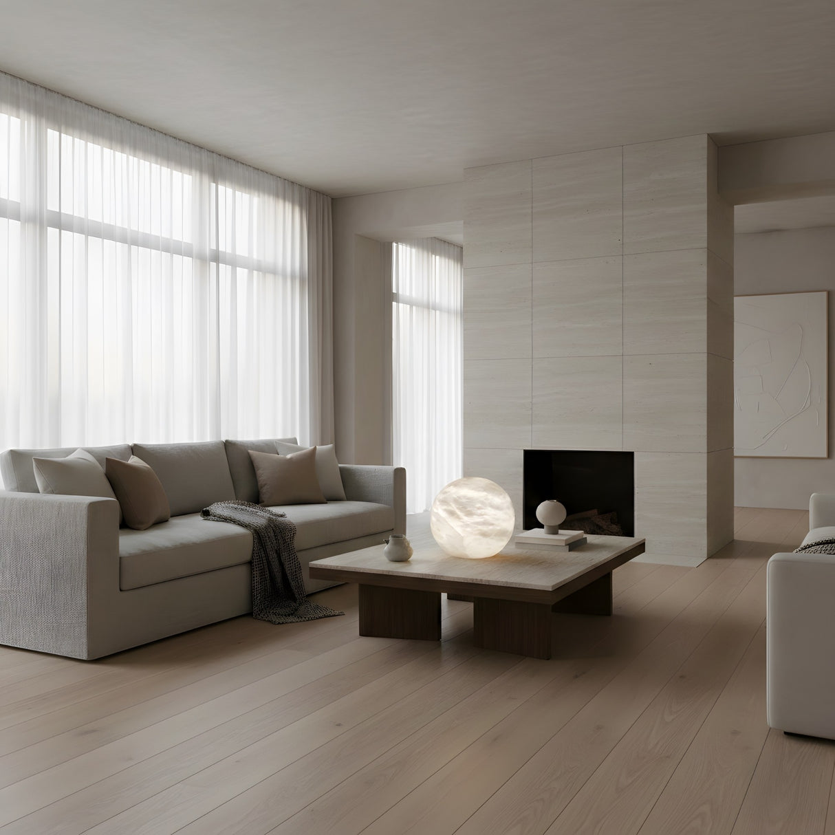 A modern, minimalistic living room with light wood floors, neutral sofas, a sleek fireplace, sheer curtains, and a low coffee table displaying the Marvello 1 Light Orb Alabaster Table Lamp - Soft White alongside candles and curated decor.