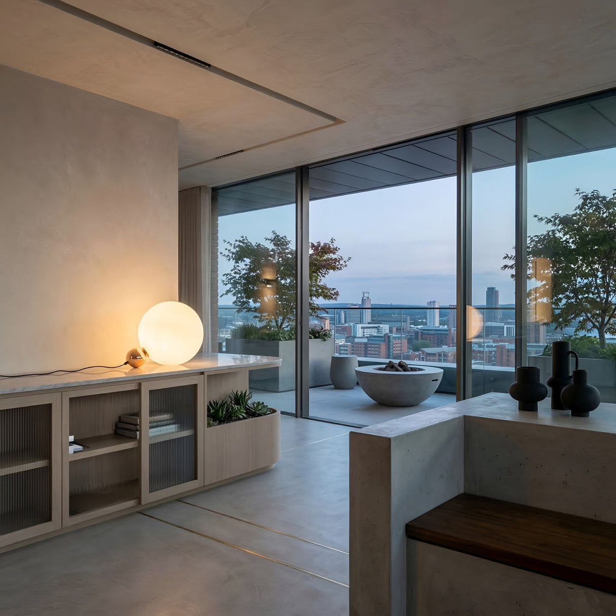 A modern living space with minimalist decor, warm lighting from the Soul Table Lamp - Gold, and glass doors opening to a balcony with potted trees and city skyline views at sunset.