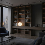 A modern living room with dark wood shelves, books, and decor features the Marvello 1 Light Orb Alabaster Table Lamp in soft white. Large windows reveal a nighttime cityscape, with dark furniture and a marble coffee table completing the look.