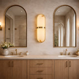 Modern bathroom with light wood double vanity, two arched mirrors, a Zantari LED Cylinder Alabaster Wall Light in brushed brass between them, gold faucets, neutral walls, and a vase of white flowers on the left side of the countertop.