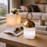 A modern living room features an Aurelio LED Ring-Handle Alabaster Table Lamp – Brass & Soft White glowing on the coffee table among books, sunglasses, and a cup, while city views shine through the window behind a cozy sofa.