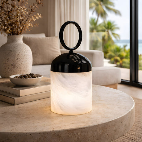 The Aurelio LED Ring-Handle Alabaster Table Lamp in matte black and soft white sits on a round beige coffee table in a bright living room with neutral decor and ocean views framed by palm trees.