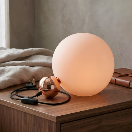 The Soul Table Lamp - Copper is a modern minimalist lamp with a glowing sphere and copper base, displayed on a wooden table alongside a folded beige blanket, closed brown notebook, and its cord and plug.
