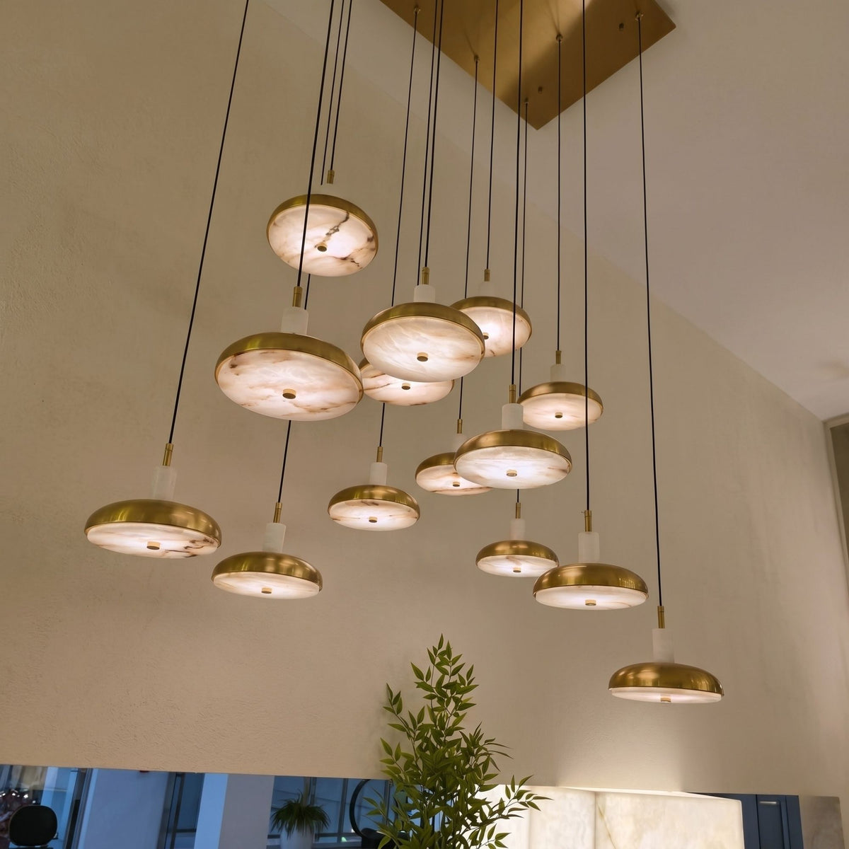 Aurevia 14 Light LED Linear Alabaster Chandelier in brushed brass, staggered domed discs suspended from a rectangular canopy, shown lit in a room with a plant.