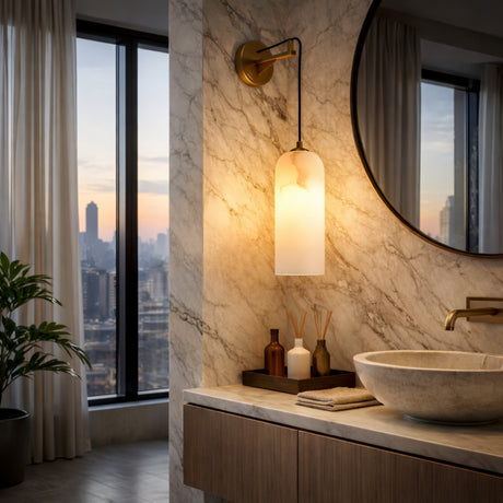 A modern bathroom with marble walls, a round mirror, a stone sink, and the Seradina 1 Light Alabaster Wall Light in soft white & brass. Large windows reveal city buildings at sunset. Decorative bottles and a plant complete the stylish look.