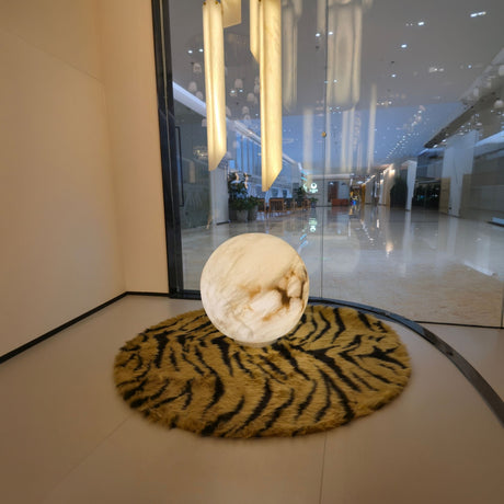 Orlena 3 Light Globe Alabaster Floor Lamp in soft white, spherical veined stone shade glowing on a tiger-print rug in a lobby setting.