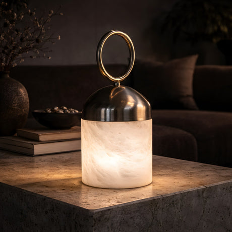 The Aurelio LED Ring-Handle Alabaster Table Lamp in Satin Nickel & Soft White glows warmly on a stone coffee table, adding modern ambient lighting to a stylish, dimly lit living room with decorative accents in the background.