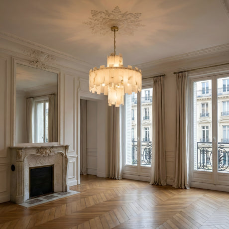 Elegant, empty Parisian room with herringbone wood floors, ornate white fireplace, tall windows with cream curtains, and a stunning Zarelli 12 Light Large Interlocking Panel Alabaster Chandelier in Soft White & Brass hanging overhead.